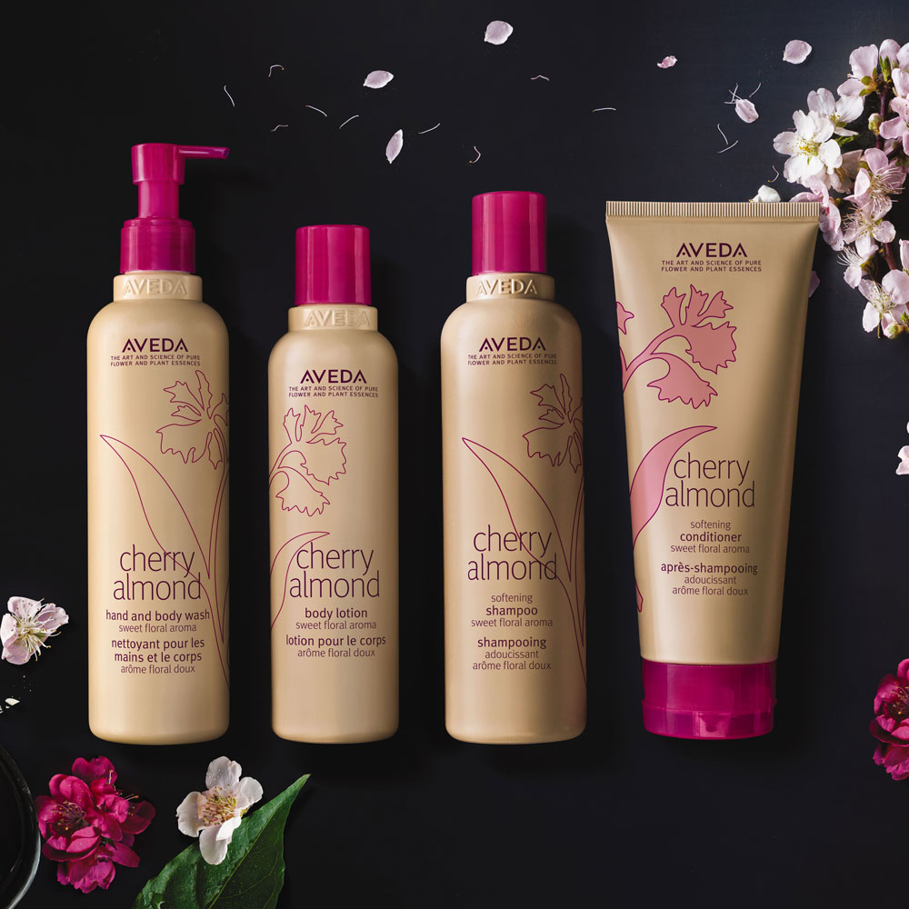 Four Aveda Cherry Almond hair and body care products are arranged on a dark surface with cherry blossoms and green leaves scattered around, evoking the ambiance of an upscale salon. - Collage Salon Day Spa | Alpharetta, GA