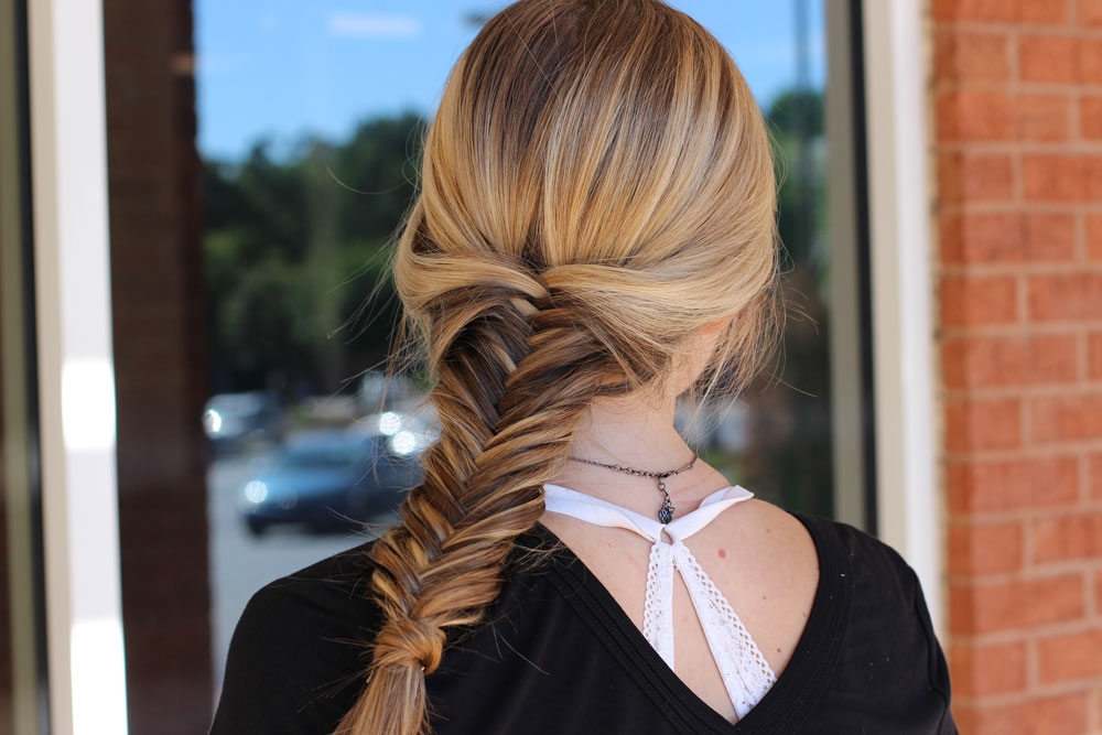 A beautiful person with long blonde hair styled in a fishtail braid, wearing a black top with a white lace insert at the neckline, stands gracefully in front of a window. - Collage Salon Day Spa | Alpharetta, GA